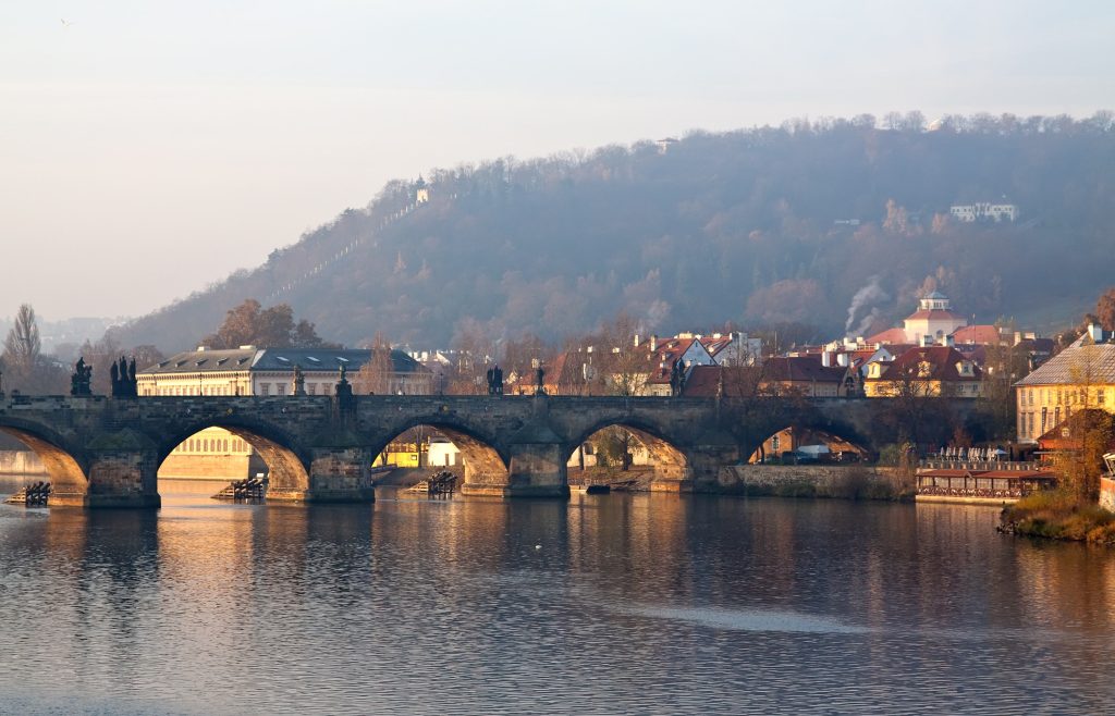 На фотографії зображено вид з води на Карлів міст у Празі. Покатий берег річки за мостом потопає у вранішньому тумані.